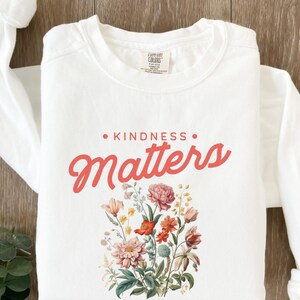 May include: White long-sleeve sweatshirt with the text "KINDNESS MATTERS" in pink lettering and a floral design in shades of pink, red, yellow, and green.