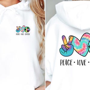May include: White hoodie with a peace sign, tie-dye heart, and soccer ball graphic. The text "Peace - Love - Soccer" is printed below the graphic.