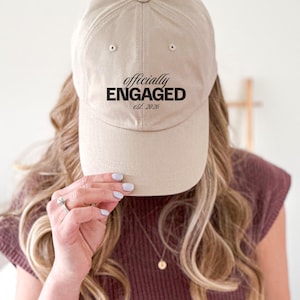 May include: A beige baseball cap with the text "officially ENGAGED est. 2026" in black. The cap is worn by a person with long blonde hair, a maroon top, and a diamond ring.