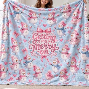 Könnte beinhalten: Eine hellblaue Fleecedecke mit einem wiederkehrenden Muster aus Schneemännern, die rosa oder blaue Wintermützen und Schals tragen, durchsetzt mit rosa Schleifen, Herzen und Schneeflocken. In der Mitte steht der rosa Text Getting my merry on.