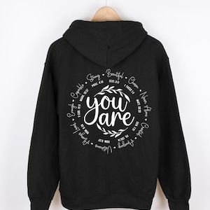 May include: Black hooded sweatshirt with a white circle graphic on the back. The graphic features the words "You are" surrounded by a wreath and a circle of words and bible verses including "Strong", "Beautiful", "Chosen", "Never Alone", "Created", "Always Loved", "Enough", "Capable".