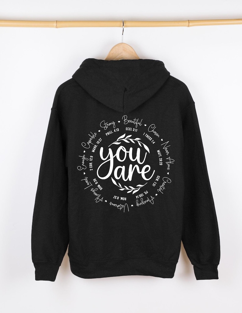 May include: Black hooded sweatshirt with a white circle graphic on the back. The graphic features the words "You are" surrounded by a wreath and a circle of words and bible verses including "Strong", "Beautiful", "Chosen", "Never Alone", "Created", "Always Loved", "Enough", "Capable".