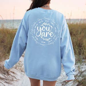 May include: Light blue sweatshirt with a white circle design on the back. The circle features the words "You are" surrounded by a wreath of leaves and a list of positive affirmations with corresponding Bible verses.
