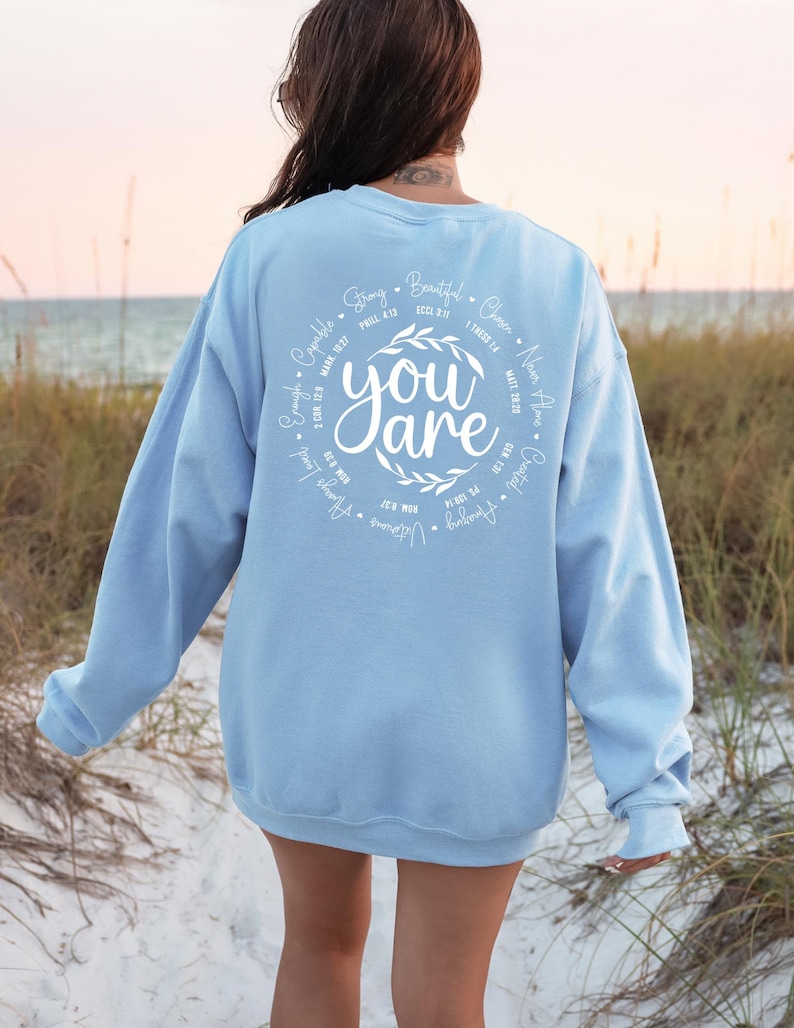 May include: Light blue sweatshirt with a white circle design on the back. The circle features the words "You are" surrounded by a wreath of leaves and a list of positive affirmations with corresponding Bible verses.