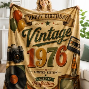以下が含まれることがあります： 「HAPPY BIRTHDAY」、「Vintage 1976」、「LIMITED EDITION」、「AGED TO Perfection」という文字がプリントされたベージュのブランケット。3つの風船、カメラ、レコード、カセットテープ、ラバランプのイラストが描かれています。