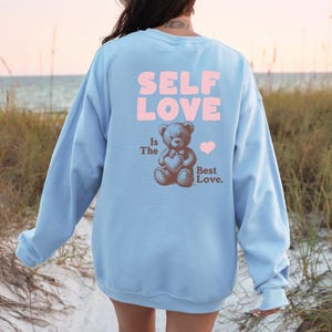 K&ouml;nnte beinhalten: Ein hellblauer Pullover mit dem Text "SELF LOVE" in rosa Buchstaben. Unter dem Text befindet sich ein brauner Teddyb&auml;r, der ein rosa Herz mit dem Text "Is The Best Love." h&auml;lt.