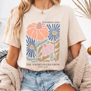 May include: A beige t-shirt with a floral design and the text "Consider How The Wildflowers Grow Luke 12:27".