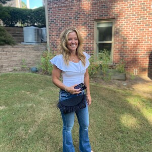 May include: A woman wearing a white ruffled top, blue jeans, and a black fringed bag. She is holding a black smartphone in her hand.