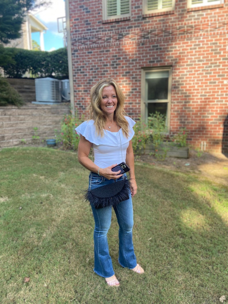 May include: A woman wearing a white ruffled top, blue jeans, and a black fringed bag. She is holding a black smartphone in her hand.