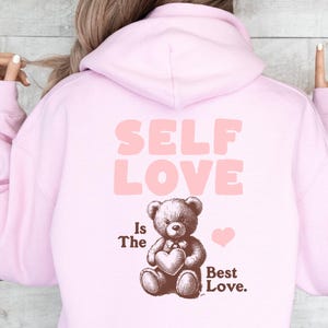 K&ouml;nnte beinhalten: Rosafarbener Hoodie mit dem Text "SELF LOVE Is The Best Love." und einem braunen Teddyb&auml;r, der ein Herz h&auml;lt.