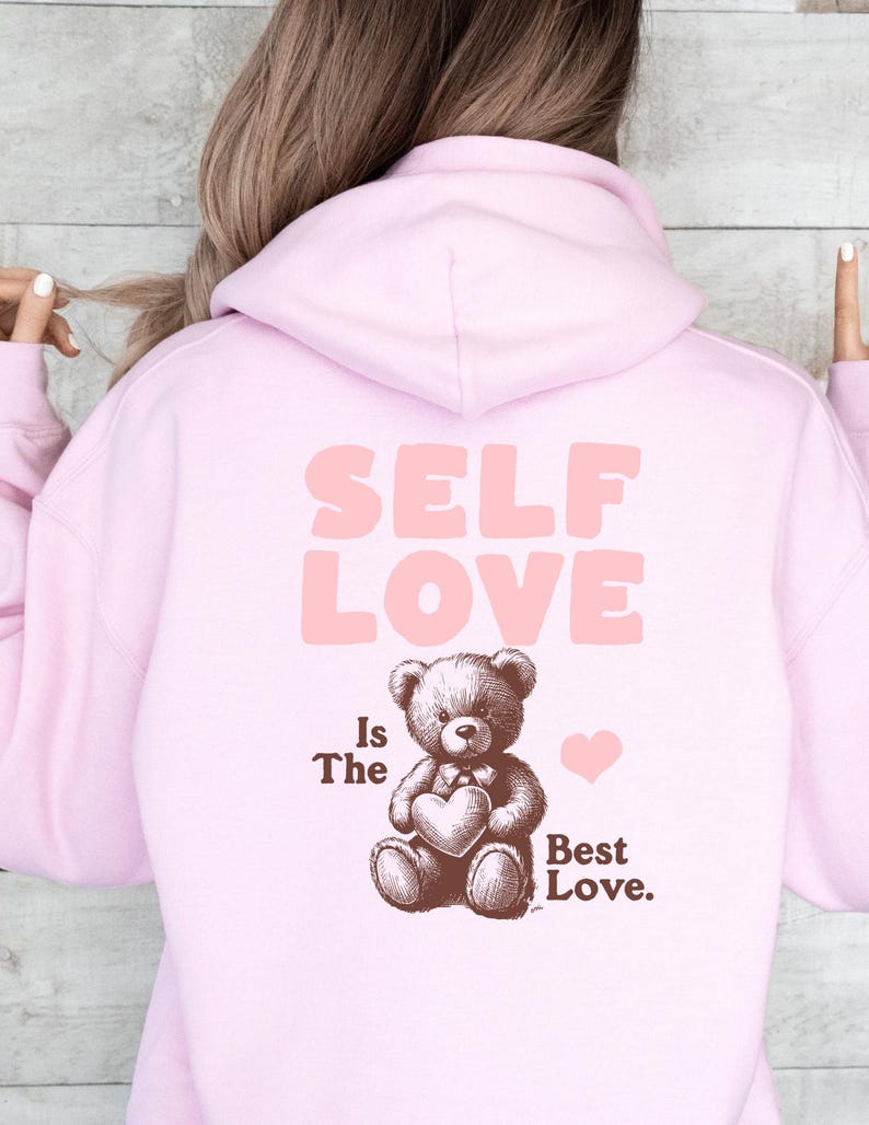 K&ouml;nnte beinhalten: Rosafarbener Hoodie mit dem Text "SELF LOVE Is The Best Love." und einem braunen Teddyb&auml;r, der ein Herz h&auml;lt.