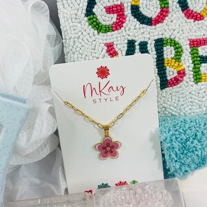 May include: A gift box filled with various items, including a gold necklace with a pink flower pendant, a white beaded pillow with the words "Good Vibes" in colorful beads, and two clear containers filled with hair scrunchies. The box also includes a white card with the words "MKay Style" printed on it.