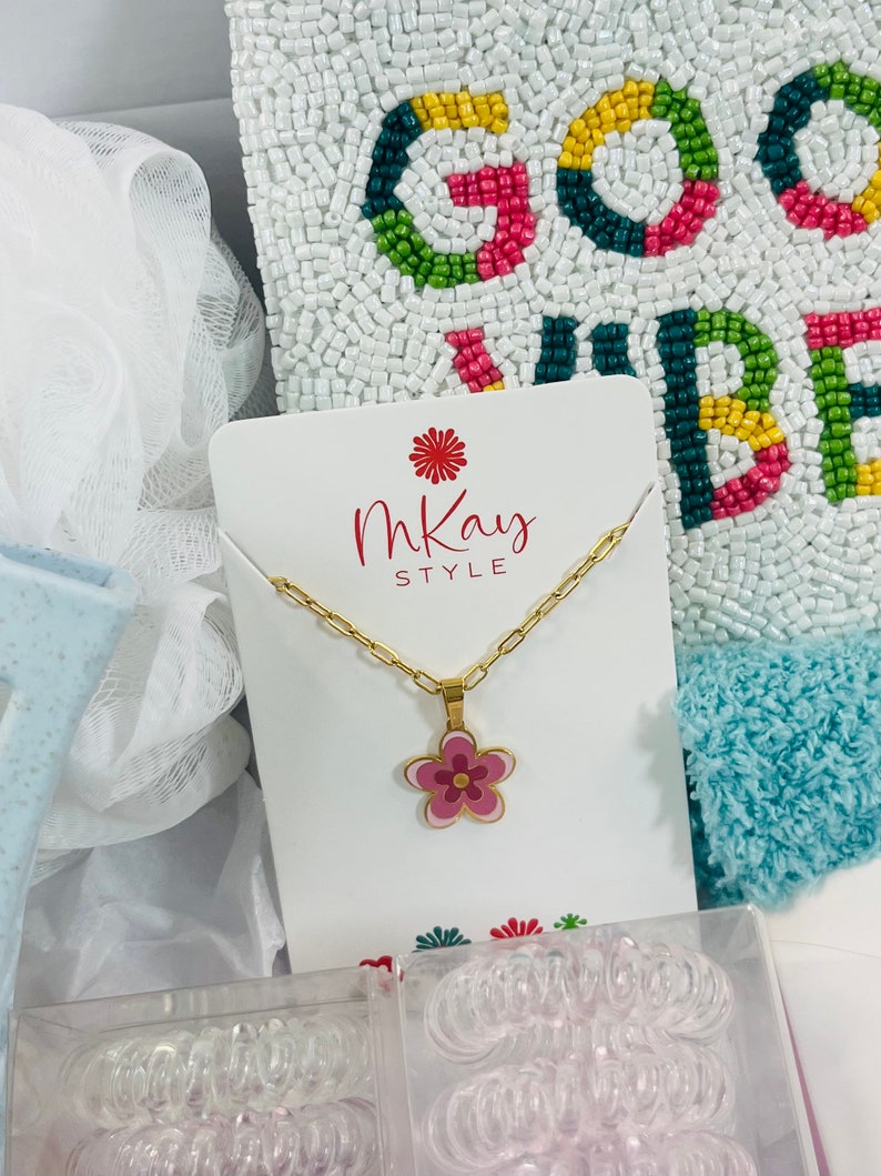 May include: A gift box filled with various items, including a gold necklace with a pink flower pendant, a white beaded pillow with the words "Good Vibes" in colorful beads, and two clear containers filled with hair scrunchies. The box also includes a white card with the words "MKay Style" printed on it.