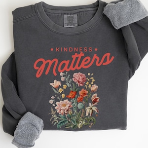 May include: A dark gray sweatshirt with the text "KINDNESS MATTERS" in red lettering. The text is surrounded by a floral design with pink, red, and yellow flowers.