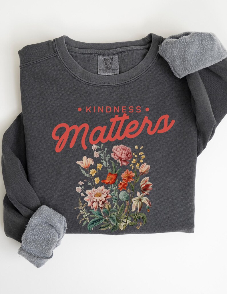 May include: A dark gray sweatshirt with the text "KINDNESS MATTERS" in red lettering. The text is surrounded by a floral design with pink, red, and yellow flowers.