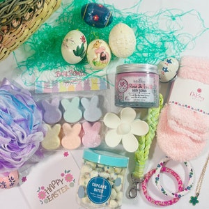 May include: A pink and green Easter gift basket with bath bombs, body scrub, a flower-shaped soap dish, a jar of cupcake bites, a pink and green beaded bracelet, a gold necklace with a shamrock charm, and a pink and white bath pouf. The basket also includes a card that says "Happy Easter" and a pink and white bath towel.