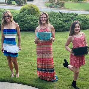 May include: Three women are standing outdoors, each holding a different style of fringe clutch purse. The purses are all in different colors and patterns. The women are wearing summer dresses.