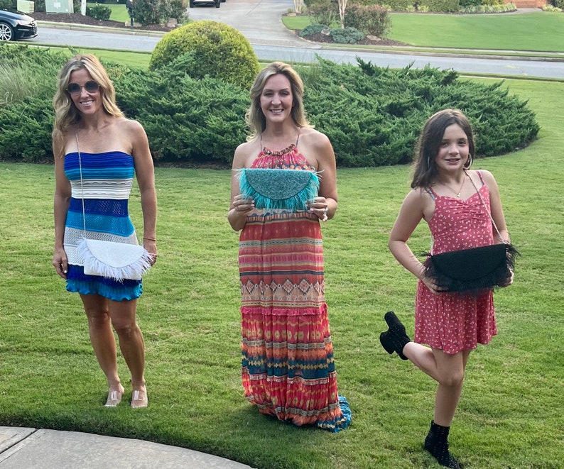 May include: Three women are standing outdoors, each holding a different style of fringe clutch purse. The purses are all in different colors and patterns. The women are wearing summer dresses.