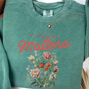 May include: Teal green sweatshirt with the text "Kindness Matters" in red lettering and a floral design.