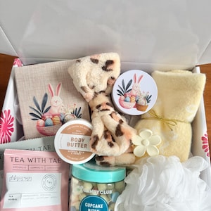 May include: An open gift box filled with self-care items. Includes a tea packet, body butter, a jar of cupcake bites, a washcloth, a loofah, and a decorative towel with an Easter bunny design. The box is white with pink floral accents.