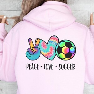 May include: Pink hoodie with a peace sign, heart, and soccer ball graphic. The graphic is in tie-dye colors. The text "Peace Love Soccer" is below the graphic.