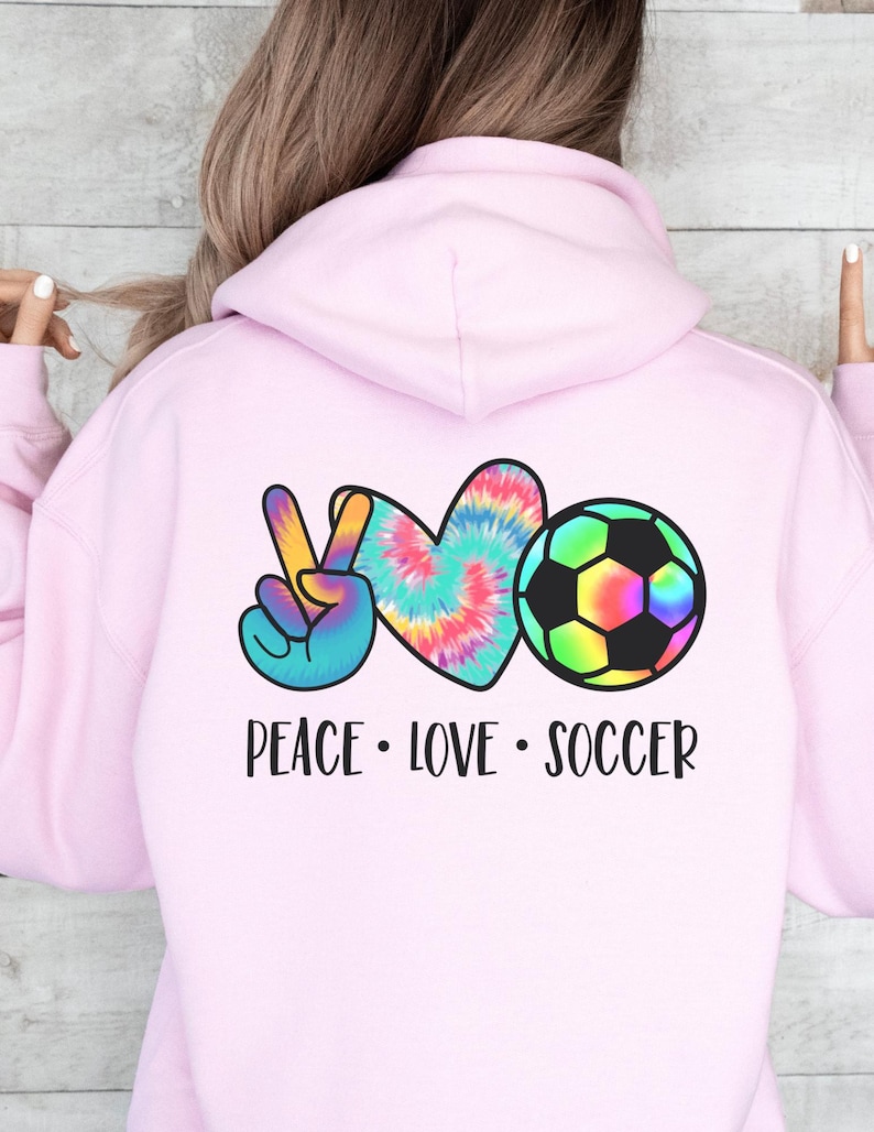 May include: Pink hoodie with a peace sign, heart, and soccer ball graphic. The graphic is in tie-dye colors. The text "Peace Love Soccer" is below the graphic.