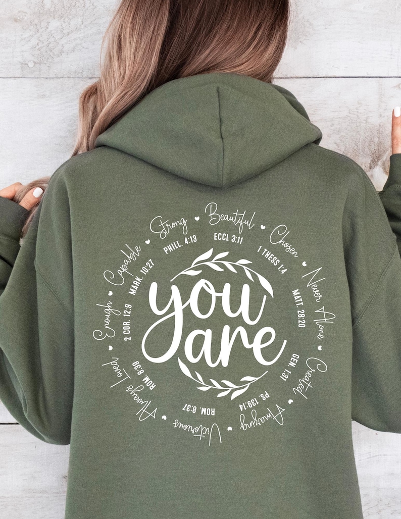 May include: A green hooded sweatshirt with a white circle design that says "you are" surrounded by inspirational words and bible verses. The words include "strong", "beautiful", "chosen", "never alone", "created", "victorious", "always love", "enough", "capable", and more.