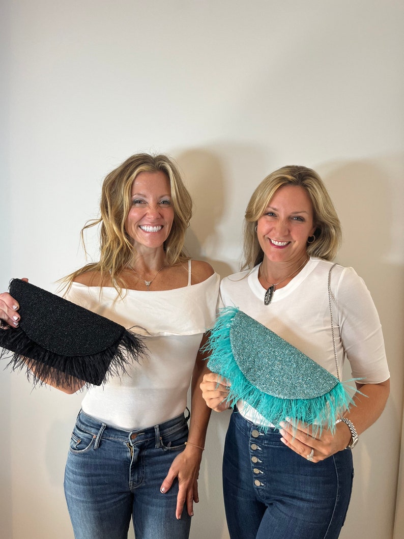 May include: Two women are holding feather-trimmed clutches. One clutch is black with black feathers, and the other is turquoise with turquoise feathers.