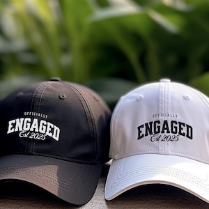 Puede incluir: Dos gorras de béisbol, una negra y una blanca, con el texto "Officially Engaged Est 2025" en la parte delantera.