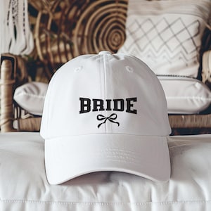 May include: A white baseball cap with the word "BRIDE" in bold, black letters and a black bow graphic. The cap is sitting on a white pillow.