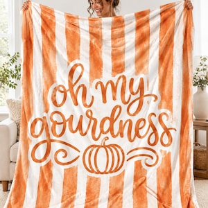 May include: A rectangular throw blanket featuring a vertical orange and white striped pattern. The center displays the orange cursive text "oh my gourdness" positioned above a simple orange line-art illustration of a pumpkin.