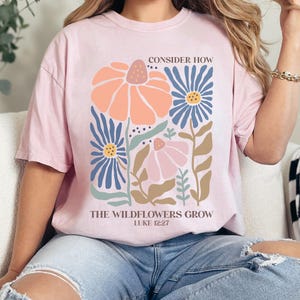 May include: A pink t-shirt with a floral design and the text "Consider how the wildflowers grow, Luke 12:27".