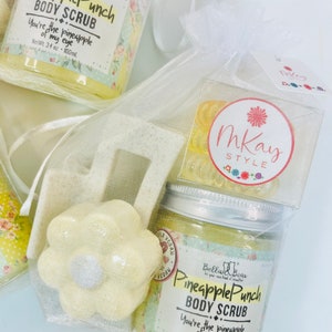 May include: A white mesh bag filled with bath products. The bag contains two jars of pineapple punch body scrub, a white bath bomb shaped like a flower, and a small tag that says "Mkay Style".