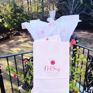 May include: A white paper gift bag with a pink flower and the text "mKay STYLE" in a circle. The bag is filled with white tissue paper.