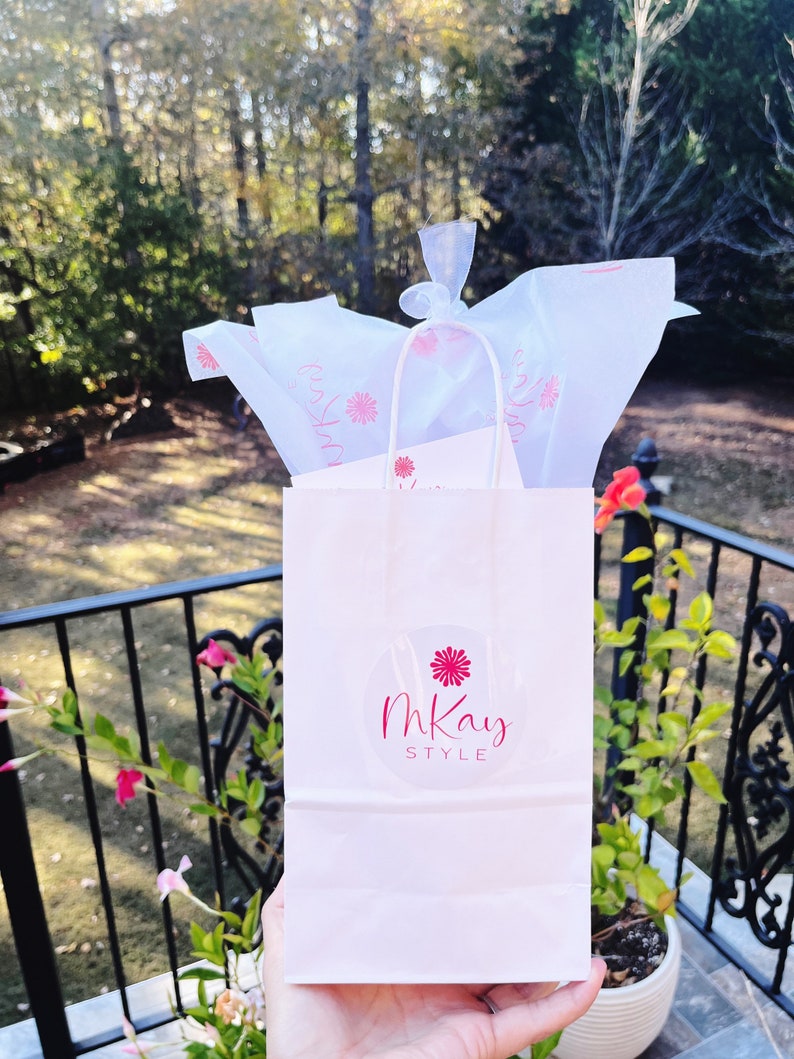 May include: A white paper gift bag with a pink flower and the text "mKay STYLE" in a circle. The bag is filled with white tissue paper.