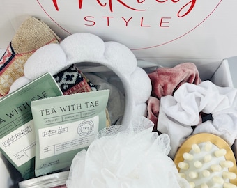 May include: A white gift box filled with self-care items, including a white headband, two pink hair scrunchies, a white bath pouf, a wooden body brush, and a jar of rose and ivy body scrub. The box also includes two tea sachets with the text "Tea with Tae" and the words "Integrity, Community, Curiosity".