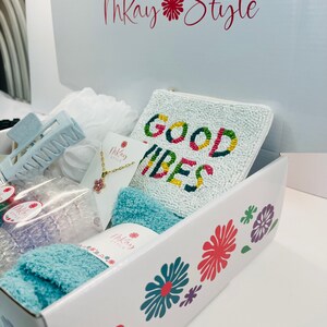 May include: A white gift box with a floral design and the text "Mkay Style" on the top. The box is open and contains a variety of items, including a blue headband, a white beaded clutch with the text "GOOD VIBES", a blue towel, and a necklace with a pink flower charm.