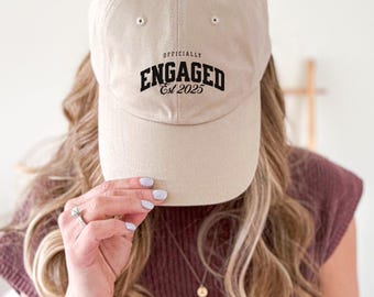 May include: A beige baseball cap with black embroidery that reads "Officially Engaged Est 2025".