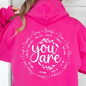 May include: Pink hoodie with a white circle graphic that says "You are" surrounded by inspirational quotes and bible verses. The quotes include "Strong", "Beautiful", "Chosen", "Never Alone", "Created", "Amazing", "Victorious", "Always Loved", "Enough", "Capable", and more.