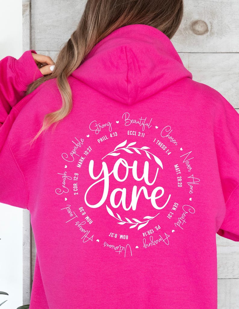 May include: Pink hoodie with a white circle graphic that says "You are" surrounded by inspirational quotes and bible verses. The quotes include "Strong", "Beautiful", "Chosen", "Never Alone", "Created", "Amazing", "Victorious", "Always Loved", "Enough", "Capable", and more.