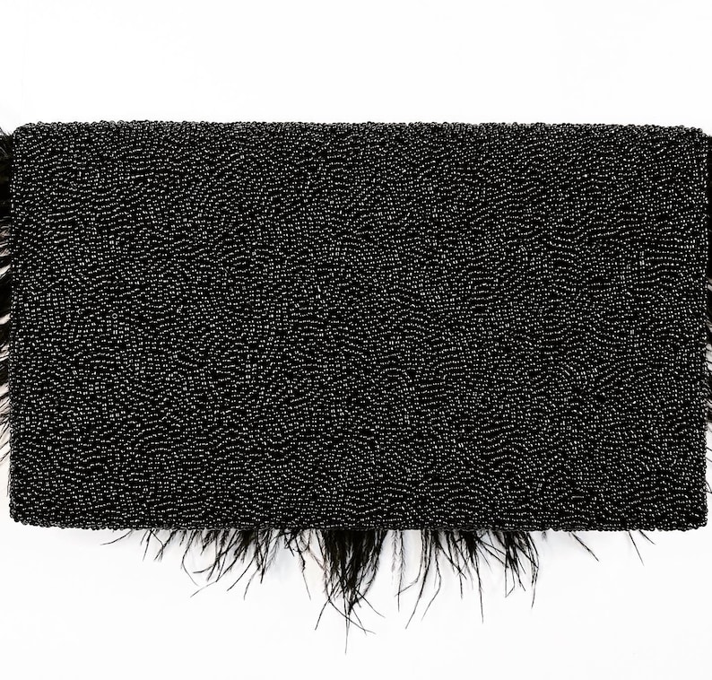 May include: A black beaded clutch with a feather trim. The clutch is made of small, tightly woven beads and has a fringe of black feathers around the bottom.