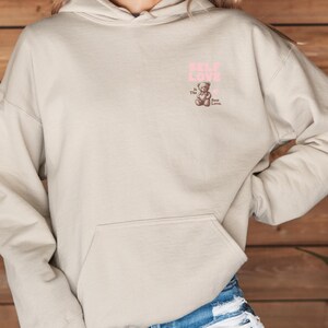 K&ouml;nnte beinhalten: Ein beiger Hoodie mit einem rosafarbenen Aufdruck eines Teddyb&auml;ren und dem Text "Self Love is the Best Love."