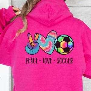 May include: Pink hoodie with a tie-dye peace sign, heart, and soccer ball graphic. The text "Peace Love Soccer" is printed below the graphic.