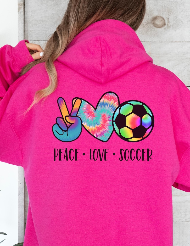 May include: Pink hoodie with a tie-dye peace sign, heart, and soccer ball graphic. The text "Peace Love Soccer" is printed below the graphic.