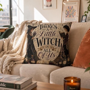 Puede incluir: Un cojín cuadrado con fondo beige y texto en negro que dice There's A Little Witch In All Of Us. El diseño incluye ilustraciones de dos figuras con sombreros de bruja, gatos negros, frascos de pociones y una escoba.