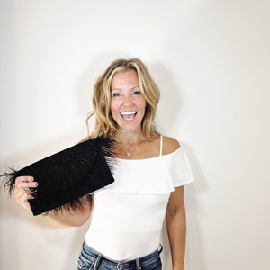 May include: A woman is holding a black clutch with a feather trim. She is wearing a white off-the-shoulder top and blue jeans.