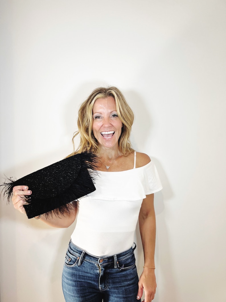 May include: A woman is holding a black clutch with a feather trim. She is wearing a white off-the-shoulder top and blue jeans.