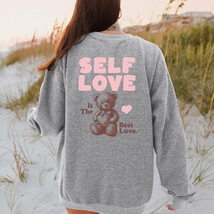 K&ouml;nnte beinhalten: Grauer Pullover mit dem Text "SELF LOVE is the Best Love." in rosa. Darunter befindet sich ein Cartoon-Teddyb&auml;r, der ein Herz h&auml;lt.