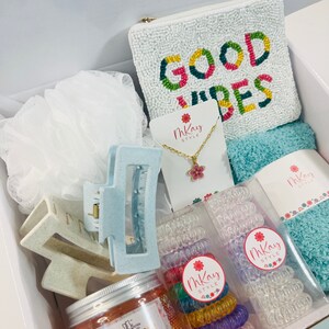 May include: A white gift box filled with various items, including a white beaded pouch with the text "GOOD VIBES" in colorful letters, a blue hair clip, a jar of orange shower jelly, a necklace with a pink flower charm, and two sets of colorful hair ties.