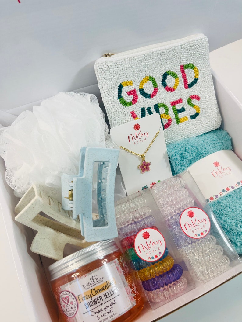 May include: A white gift box filled with various items, including a white beaded pouch with the text "GOOD VIBES" in colorful letters, a blue hair clip, a jar of orange shower jelly, a necklace with a pink flower charm, and two sets of colorful hair ties.